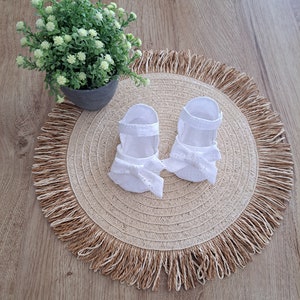 May include: A pair of white baby sandals with eyelet detailing and bow accents, resting on a round, woven placemat with a fringed edge. A small potted plant with green foliage sits nearby on a wooden surface.
