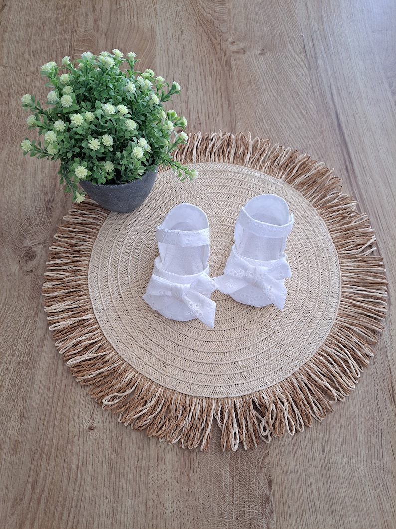 May include: A pair of white baby sandals with eyelet detailing and bow accents, resting on a round, woven placemat with a fringed edge. A small potted plant with green foliage sits nearby on a wooden surface.