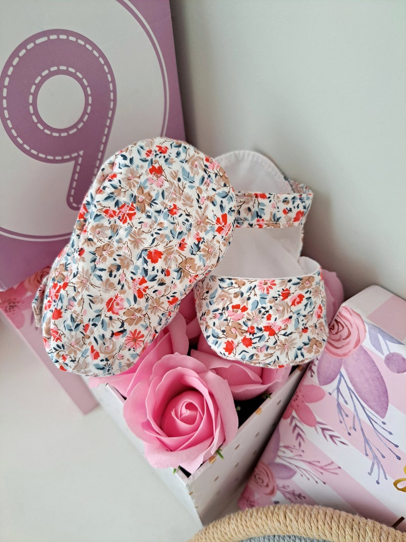 May include: Baby sandals with a floral pattern in red, blue, and brown on a white background. The sandals are placed in a decorative box with a pink rose. A large number 9 is visible in the background.