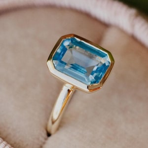 May include: A gold ring with a light blue emerald-cut gemstone. The ring is sitting in a light pink ring box.