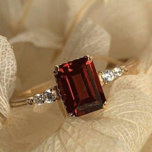 Op de afbeelding: Een gouden ring met een grote, donkerrode, smaragd geslepen edelsteen en kleinere, heldere edelstenen op de band.