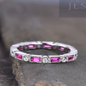 Puede incluir: Un anillo de plata con piedras rosas rectangulares alternas y pequeñas piedras claras redondas. El anillo está sobre una superficie gris oscuro texturizada. El anillo es una banda de eternidad completa.