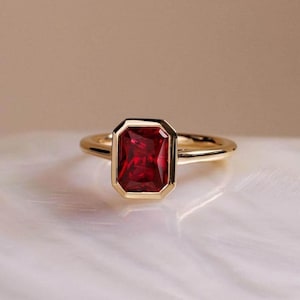 May include: A gold ring with a red emerald-cut gemstone set in a bezel setting.
