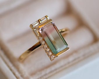 May include: A gold ring featuring a rectangular, multi-colored gemstone with pink, green, and clear sections. The ring has a delicate band and is accented with small, clear stones and rectangular accents on the top.