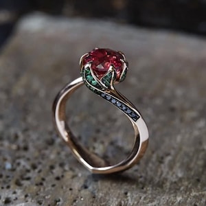 May include: A rose gold ring with a large, round, red gemstone. The ring's design includes green and black accent stones, and a distinctive, flowing band. The ring is set against a textured, grey background.