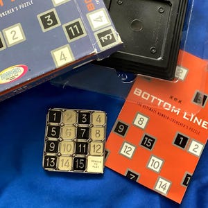 May include: A Bottom Line number puzzle game set including a blue cardboard box with white and black numbered tiles, a clear plastic tray, a black square base, and an orange instruction card. The metal puzzle features a 4x4 grid of numbers.