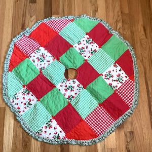 May include: A round Christmas tree skirt with a patchwork design. The skirt features red, green, and white squares with gingham and floral patterns. The edge is trimmed with a green and white crocheted border. The skirt has a hole in the center.