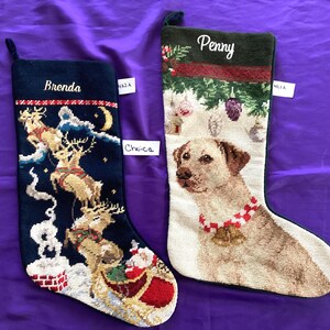 May include: Two needlepoint Christmas stockings. The left stocking is dark blue with a scene of Santa in a sleigh pulled by reindeer and the name Brenda. The right stocking is cream with a Labrador dog wearing a bell collar and the name Penny.
