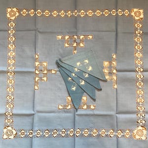 May include: A blue linen tablecloth with a white lace border and four blue linen napkins. The tablecloth has a geometric pattern in the center.