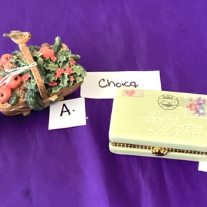 May include: Two decorative items on a purple surface. One is a basket with a bird on top, filled with red apples and greenery. The other is a light green trinket box with a gold clasp and a postage stamp design.
