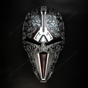 Op de afbeelding: Een gedetailleerd zilver en bruin masker met ingewikkelde bloempatronen. Het masker heeft een centraal verticaal ontwerp en een puntig ondergedeelte. De achtergrond is zwart, wat de metalen uitstraling van het masker benadrukt.