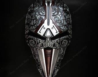 Op de afbeelding: Een gedetailleerd zilver en bruin masker met ingewikkelde bloempatronen. Het masker heeft een centraal verticaal ontwerp en een puntig ondergedeelte. De achtergrond is zwart, wat de metalen uitstraling van het masker benadrukt.