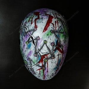 Pode incluir: Uma máscara branca desgastada com uma superfície pintada e texturizada. A máscara apresenta respingos de tinta vermelha, roxa, verde e azul, com um efeito de sangue. Possui um design costurado com arame e detalhes coloridos.
