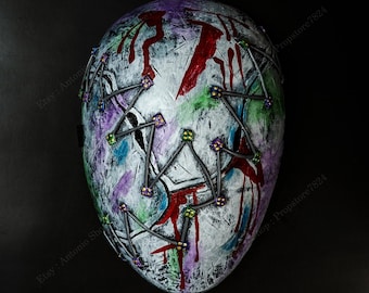 May include: A distressed, white mask with a painted, textured surface. The mask features red, purple, green, and blue paint splatters, with a blood-like effect. It has a stitched design with metal wire and colorful accents.