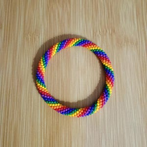 May include: A vibrant, rainbow-colored beaded bracelet. The bracelet is a continuous loop of small, colorful beads in a spectrum of hues, including red, orange, yellow, green, blue, and purple. The bracelet is displayed on a light brown wooden surface.
