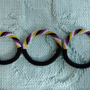 May include: Three beaded bracelets in black, white, yellow, and purple. The bracelets are arranged in a circle on a light blue knitted fabric.