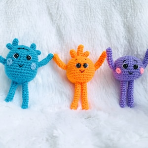 May include: Three crocheted plush toys with smiling faces. The toys are blue, orange, and purple and have long arms and legs.