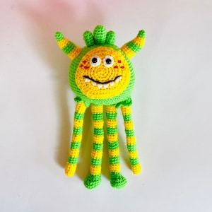 Puede incluir: Un juguete monstruo de crochet verde y amarillo con una cara sonriente y seis patas.
