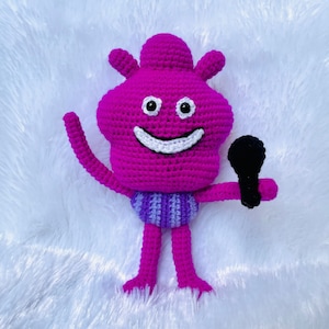 Puede incluir: Un juguete de peluche rosa tejido a crochet con una cara sonriente, que lleva un atuendo a rayas morado y blanco y sostiene un micrófono negro.