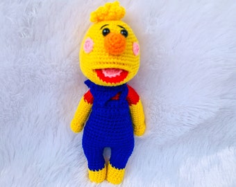 May include: A crocheted yellow bird doll with a red mouth, orange nose, and pink cheeks. The doll is wearing blue overalls with red accents.