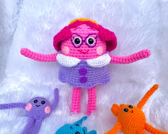May include: A collection of handmade crochet dolls in various colors. The main doll is pink with a purple dress, white collar, and glasses. Other dolls are purple, blue, and orange. The dolls have outstretched arms and are set against a white, textured background.