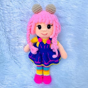 Peut inclure: Une poupée en crochet avec des cheveux roses, des vêtements bleus et jaunes et des chaussures roses. La poupée est assise sur une surface bleu clair et duveteuse.