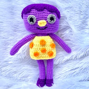 May include: A crocheted purple doll with a yellow and orange polka dot dress. The doll has large eyes and a small nose.