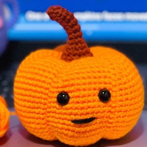 May include: A crocheted orange pumpkin with a brown stem and black eyes and a mouth. The pumpkin is sitting on a gray surface.