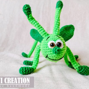 May include: A green crocheted toy with large eyes and a smiling mouth. The toy has long arms and legs and is made in a whimsical style.