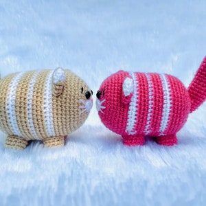 May include: Two crocheted cat plushies facing each other. The cat on the left is tan with white stripes and the cat on the right is pink with white stripes.
