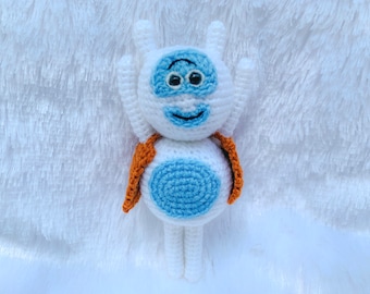 May include: A white crocheted plush toy with blue eyes, a blue mouth, and a blue circle on its chest. The toy is wearing a brown crocheted cape.
