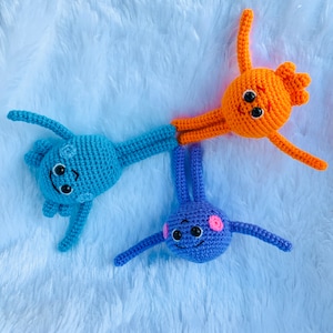 May include: Three crocheted stuffed toys with smiling faces. One is blue, one is orange, and one is purple.