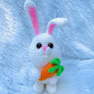 Könnte beinhalten: Ein weißer Häkel-Kaninchen, der eine kleine orange Karotte mit grünen Blättern hält. Das Kaninchen hat rosa Ohren und braune Augen.