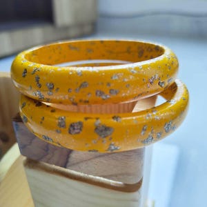 May include: Two yellow bangles with silver flecks are stacked on top of each other. The bracelets are thick and rounded, with a glossy finish. The background is blurred, suggesting a focus on the jewelry.