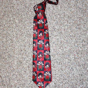 May include: A necktie featuring a repeating checkered pattern of red and dark blue squares. The red squares contain oval frames with illustrations of Mickey Mouse and Donald Duck wearing Santa hats, surrounded by green holly and red berries.