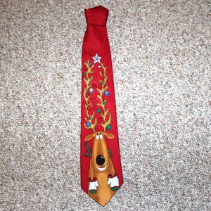 May include: A red necktie featuring a cartoon reindeer with large antlers decorated with colorful Christmas ornaments and a white star at the top. The reindeer has a brown face, a large black nose, and is wearing white mittens with green cuffs.