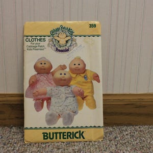 May include: A vintage Butterick pattern for Cabbage Patch Kids premie clothes. The pattern features illustrations of three Cabbage Patch Kids dolls wearing different outfits. The pattern is in a yellow and green color scheme.