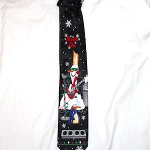May include: A black necktie featuring a white snowflake pattern and a graphic of a snowman with a mohawk playing a red electric guitar. The snowman wears green sunglasses and a black leather jacket. Below, a penguin stands near a string of lights.