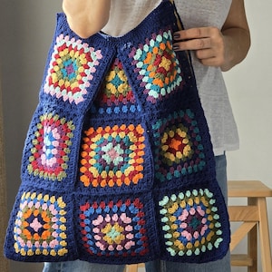 Könnte beinhalten: Eine große, handgefertigte Tragetasche mit dunkelblauem Hintergrund und bunten Häkelquadraten. Die Quadrate weisen eine Vielzahl von Farben auf, darunter Rosa, Gelb, Orange und Grün. Die Tasche hat einen braunen Lederriemen.