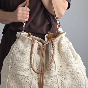 Könnte beinhalten: Cremefarbene, große Häkeltasche mit Kordelzugverschluss und Lederakzenten. Die Tasche hat ein geometrisches Muster und geflochtene Ledergriffe. Die Tasche hat einen hellbraunen Leder-Schulterriemen.
