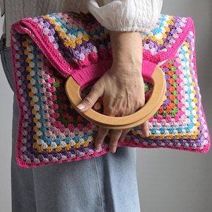 May include: A colorful, handmade crochet clutch with a wooden handle. The bag features a granny square design in various colors, including pink, purple, yellow, and blue. The handle is a light brown, and the bag is held by a person.