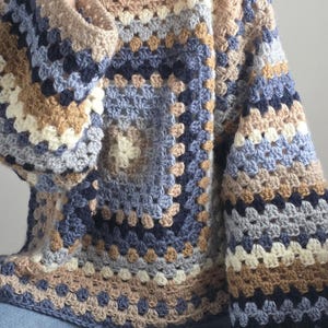 Könnte beinhalten: Ein langärmeliger Pullover aus gehäkeltem Garn in einem Granny-Square-Muster. Das Kleidungsstück weist eine Farbpalette aus Marineblau, Hellblau, Beige, Creme und Grau auf, die in konzentrischen quadratischen Reihen angeordnet sind.