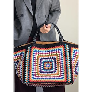 Puede incluir: Un bolso tote grande y colorido de ganchillo con un patrón cuadrado en varios colores, incluyendo azul, rojo, amarillo y morado. El bolso tiene ribetes negros y asas de cuero. El bolso es sostenido por una persona que lleva una blazer gris.