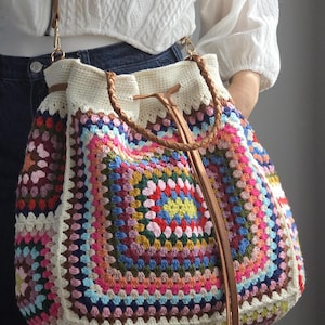 Può includere: Una grande borsa all'uncinetto colorata con un design a granny square. La borsa presenta una parte superiore color crema e un cinturino in pelle marrone. Il corpo della borsa è un patchwork di colori vivaci, tra cui rosa, blu, giallo e verde.