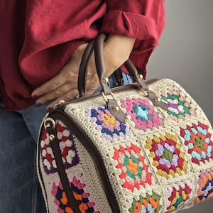 May include: A beige crocheted handbag with colourful granny squares and brown leather accents. The bag has two top handles and a zip closure.