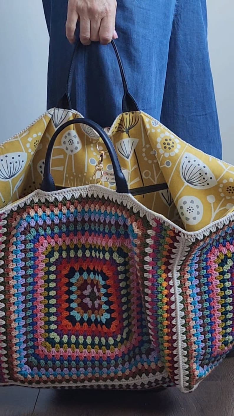 May include: A large, colorful crochet tote bag with a yellow floral fabric lining. The bag has a black leather handle and a zipper closure.