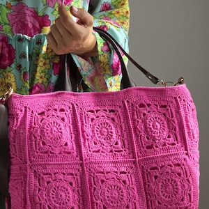 Könnte beinhalten: Eine handgefertigte, leuchtend rosa Häkeltasche mit quadratischem Muster. Die Tasche hat dunkelbraune Ledergriffe und einen Schulterriemen. Die Tasche wird von einer Person getragen, die ein Blumenkleid trägt.