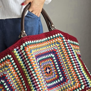 Puede incluir: Un bolso tote grande y colorido de ganchillo con un diseño de patchwork. El bolso presenta un borde burdeos y asas de cuero marrón. El patrón de ganchillo incorpora varios colores, creando un accesorio vibrante.