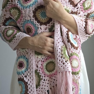 May include: A light pink crocheted shawl with a floral pattern in shades of pink, brown, green, and blue. The shawl has a fringe along the bottom edge. The shawl is worn over a white top.
