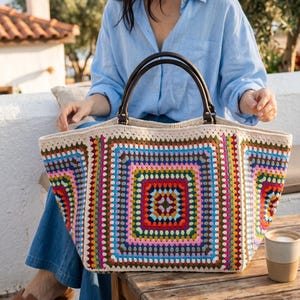 May include: A large, rectangular crochet tote bag featuring a vibrant granny square pattern in multiple colors including pink, blue, green, red, and yellow, framed by cream-colored yarn borders, with two dark brown leather handles attached by metal rings.
